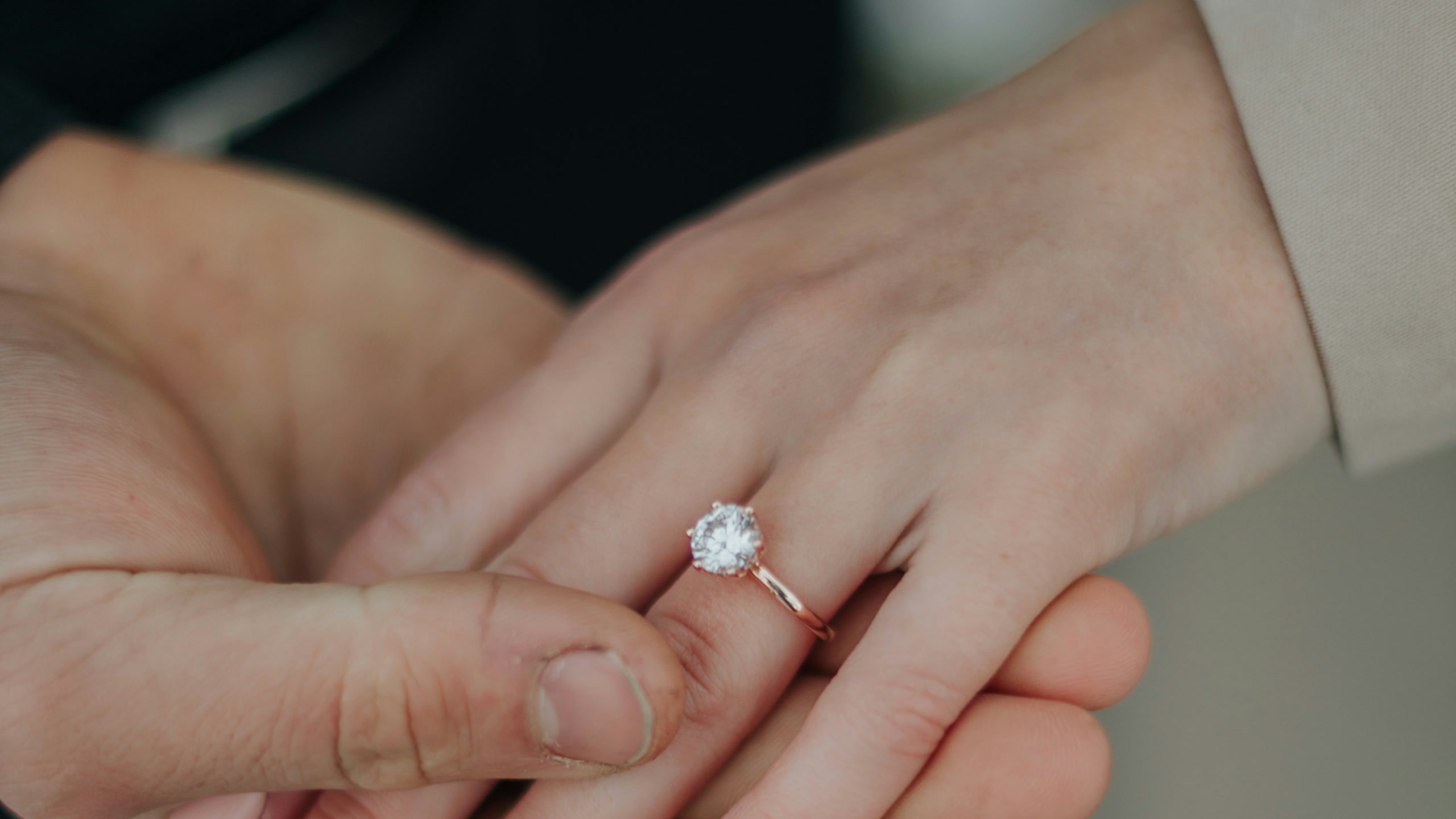Two hands rest together, a solitaire diamond ring on her ring finger.